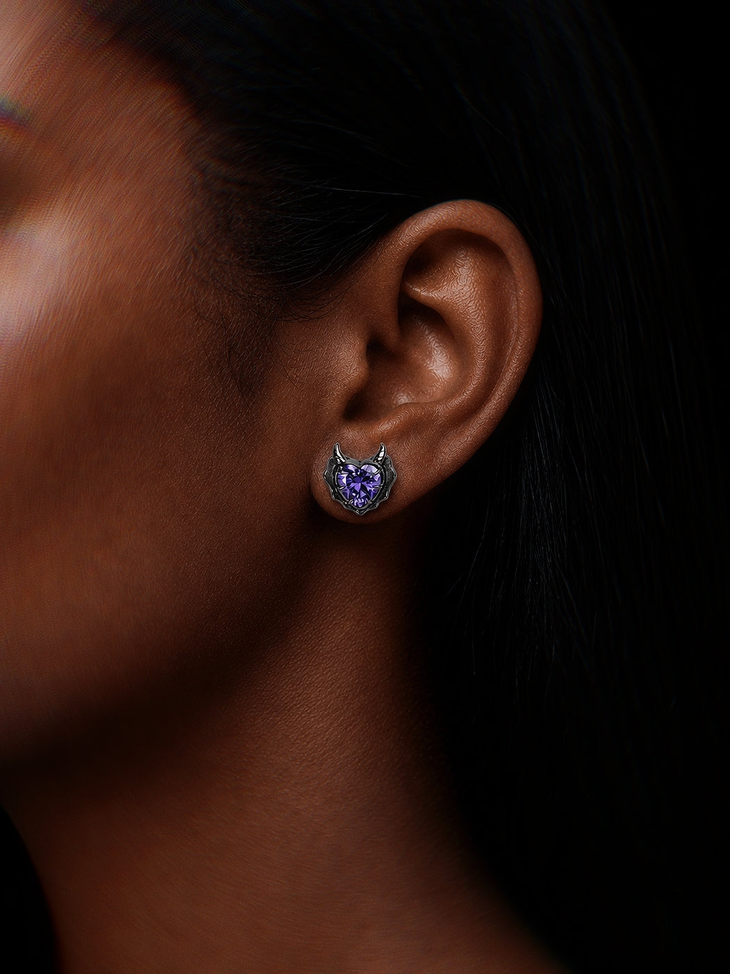 A close-up side profile of a woman's ear wearing a small devil-themed stud earring with a purple heart-shaped gemstone and silver horns, set against a dark background.